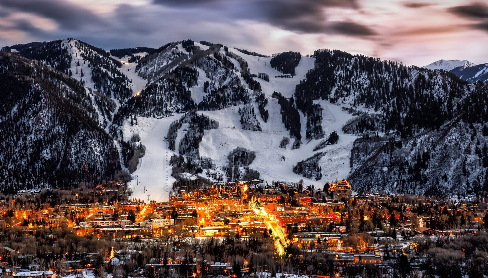 Ski runs cut through forested slopes above the lit town at dusk